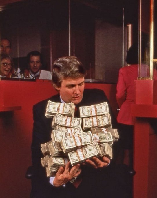 A vintage-style photograph of a man in a dark suit holding a massive pile of banded stacks of US currency in his arms.