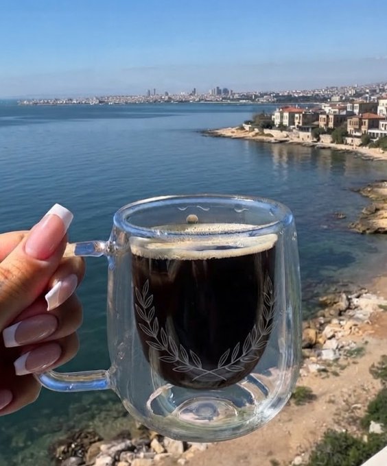 A hand holds a double-walled glass mug of black coffee, overlooking a coastal city and sea from a rocky vantage point.