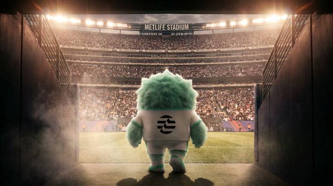 A green-furred mascot wearing a jacket featuring an onchain logo walks out of a tunnel onto the field at MetLife Stadium, with the stadium name and "2026 FIFA World Cup" text visible above the packed stands, visually echoing the post's message by merging global sports spectacle with onchain culture.