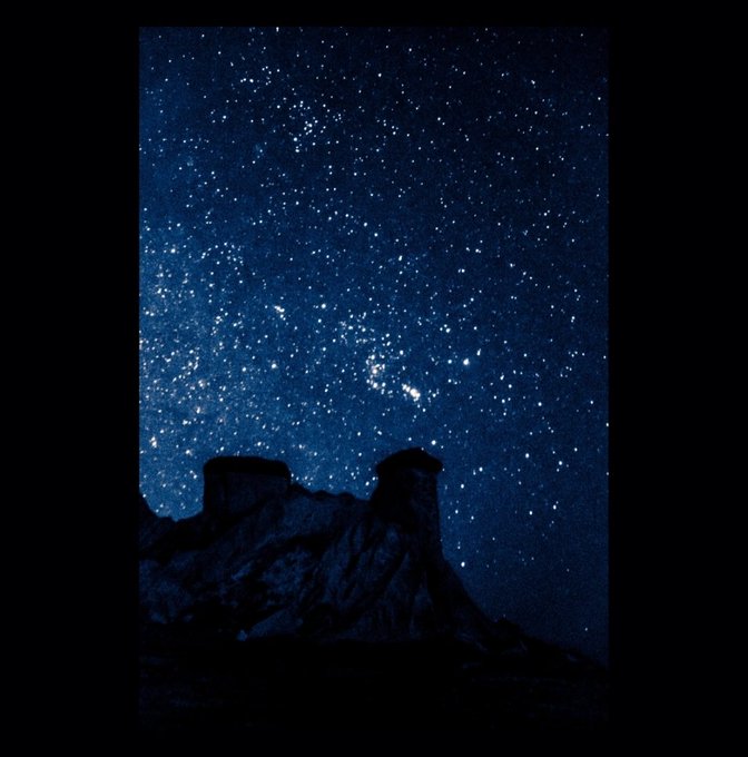 A starry night sky filled with countless stars and the visible band of the Milky Way stretches above the dark, silhouetted ridges and hoodoos of a desert landscape.