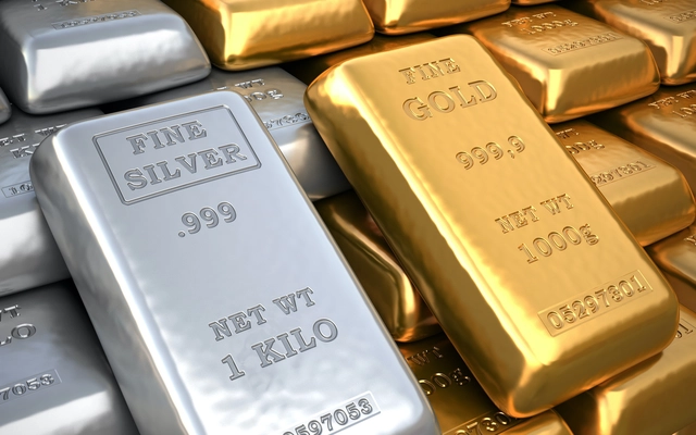 A close-up photograph showing stacked bullion bars, including a prominent silver bar labeled "FINE SILVER .999 NET WT 1 KILO" and gold bars labeled "FINE GOLD 999.9 NET WT 1000g".