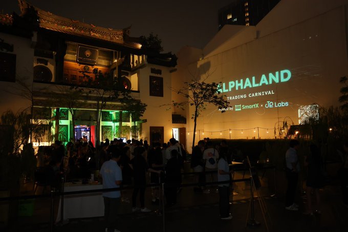 A nighttime outdoor event at 'ALPHALAND TRADING CARNIVAL' sponsored by Mantle, SmartX, and JP Labs, showing a crowd of attendees gathered near a building with traditional Chinese architecture on the left.