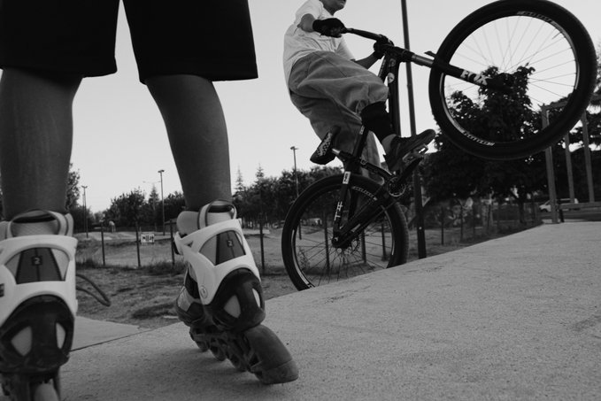 A black and white photo showing a close-up of someone's legs in inline skates in the foreground, with a child riding a BMX bike performing a wheelie in the background.