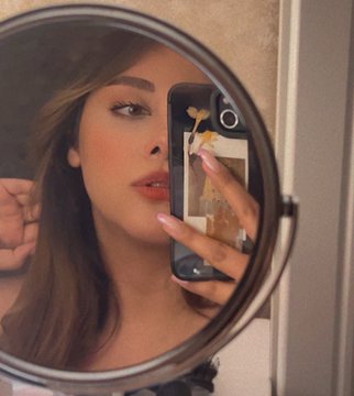 A mirror selfie of a woman holding a smartphone, capturing her reflection in a round vanity mirror.