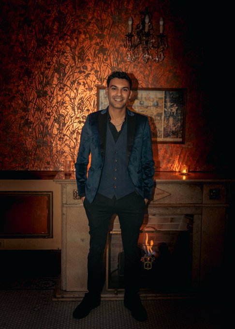 A smiling young man in a blue blazer and vest posing in front of a fireplace with a fire, beneath an ornate chandelier and patterned orange wallpaper.