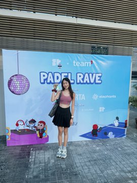 A woman in a pink top and black skirt poses in front of a "PADEL RAVE" banner featuring "team" and "SuperteamTH" branding, holding up a small item while standing on a paved outdoor area. The image captures her attendance at the Superteam Thailand Padel Rave event, aligning with the post's narrative of her venturing into the web3 space as a newcomer and engaging with the community at this social gathering.