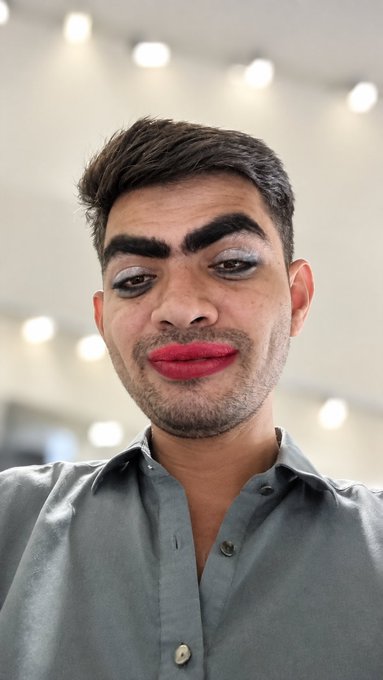 A man with dark hair, dramatic eye makeup, and bright pink lipstick poses for a selfie in a gray shirt.