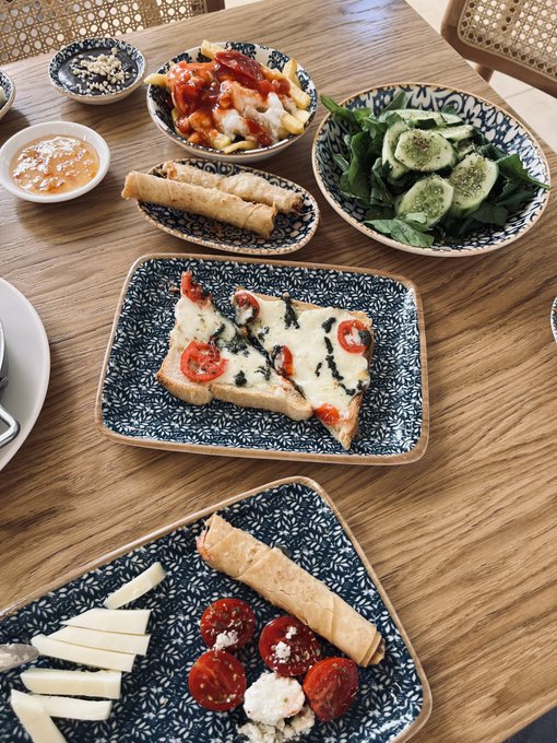 An overhead view of a wooden table set with various dishes including a flatbread with tomatoes and cheese, a cucumber salad, spring rolls, and dipping sauces.