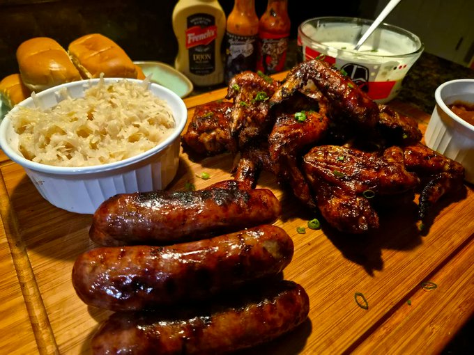 A wooden board displaying grilled sausages, BBQ ribs, sauerkraut in a white bowl, dinner rolls, French's mustard, hot sauce bottles, and ranch dressing.