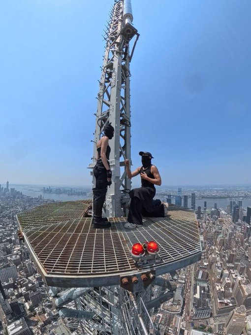 A high-angle photograph showing two people standing and kneeling on a small metal platform at the very top of a tall communications spire, with a city skyline and water visible far below under a clear blue sky.