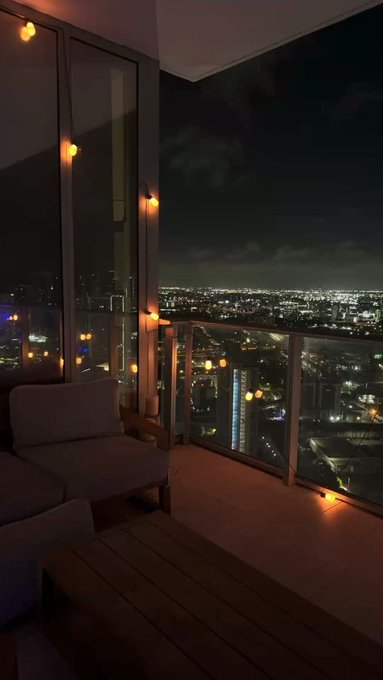 A nighttime photo taken from a high-rise balcony featuring string lights along the railing, a white outdoor sofa, and a panoramic view of a brightly lit city skyline with dramatic clouds above.