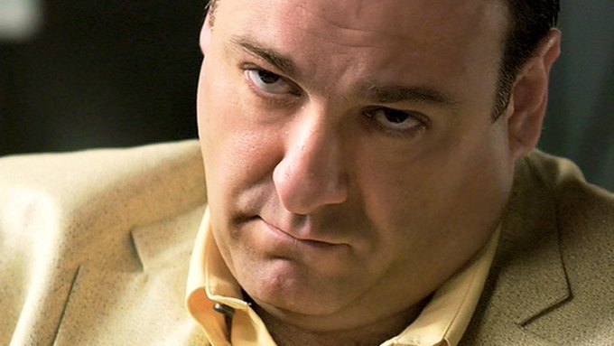 A close-up portrait of a man with dark hair wearing a tan jacket and orange shirt, looking downward.
