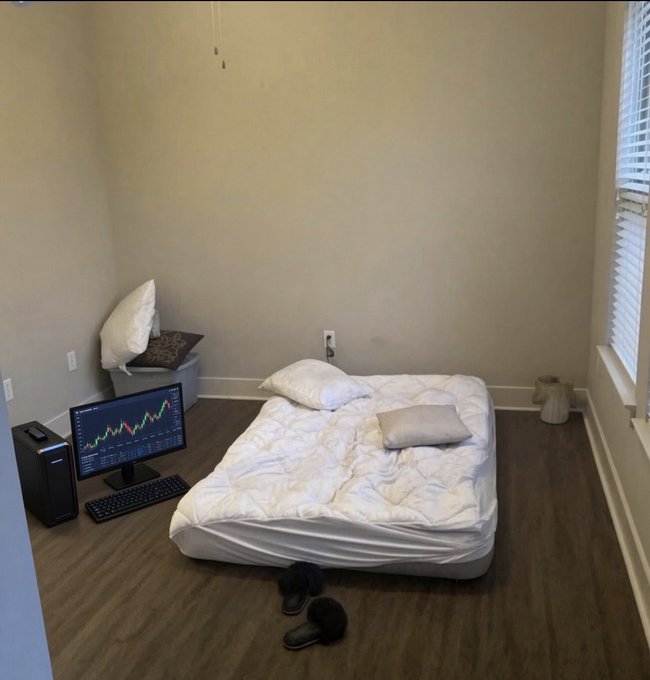 A minimalist bedroom with a mattress on the floor, a computer setup displaying a stock chart, and a small trash bin in the corner.
