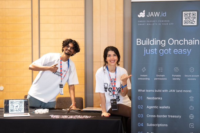 Two people in white t-shirts posing behind a booth for 'JAW.id' (a smart wallet app) with a banner reading 'Building Onchain just got easy', listing features like 'Instant onboarding', 'Onchain permissions', 'Portable identity', and 'Secure account recovery'.