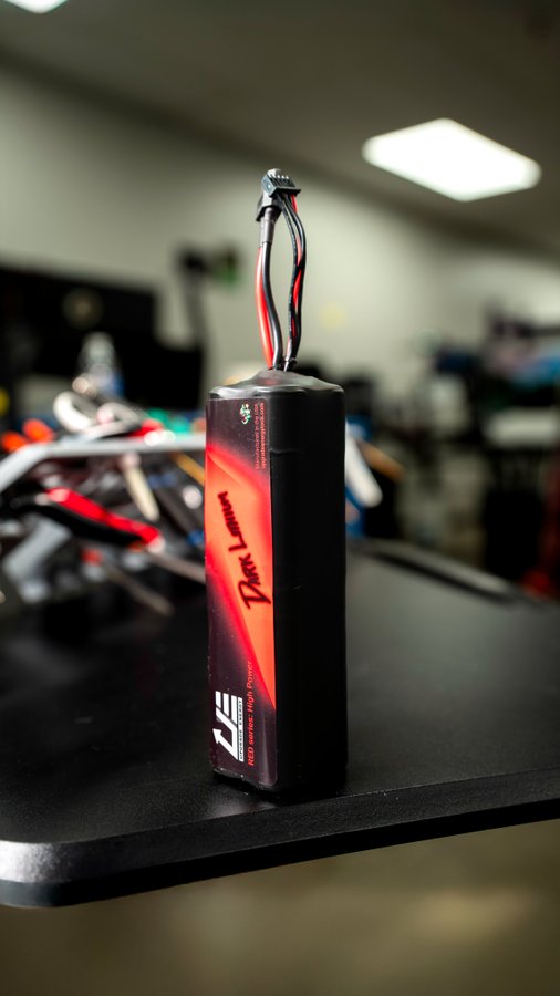 A rectangular electronic module stands upright on a black lab bench, with red, black, and blue wires protruding from the top. Its label includes “ZE” and “2200 mAh,” while the remaining small text is not clearly readable.