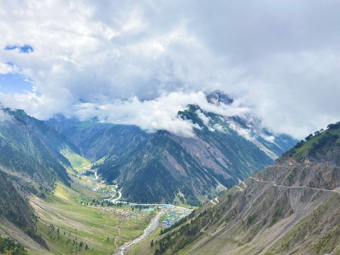 A scenic mountain valley landscape with green slopes, a winding river through a small settlement, and low-hanging clouds partially obscuring the peaks.