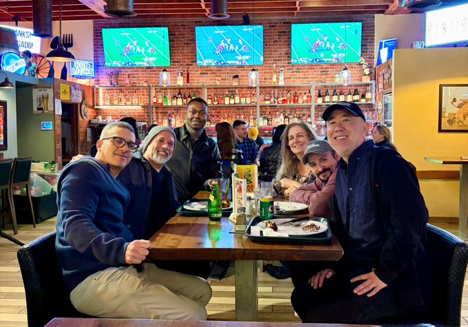 A group of seven people pose together smiling around a table at a restaurant or bar, with TVs showing sports in the background.