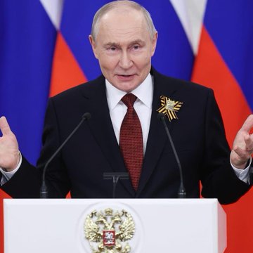 Russian President Vladimir Putin stands at a podium adorned with the Russian coat of arms, wearing a dark suit with a red tie and a gold ribbon pin, gesturing with both hands raised as he speaks, with Russian flags behind him.