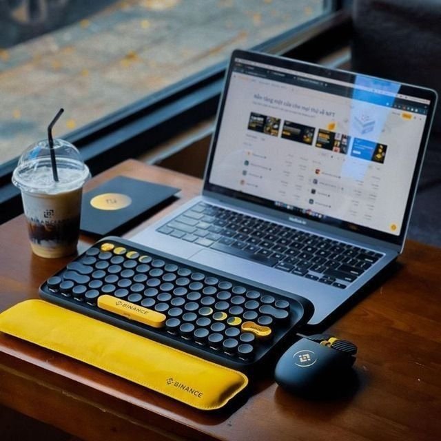 A workspace setup featuring a MacBook displaying the Binance website alongside a yellow and black Binance-branded mechanical keyboard and matching wrist rest, plus an iced coffee on a wooden table.