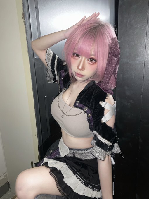 A young woman with short pink hair poses indoors, wearing a black velvet and lace outfit with a chain necklace.
