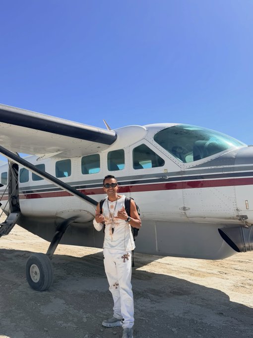 Evan, dressed in a crisp white outfit and sunglasses, stands confidently beside a small Cessna aircraft on a sandy airstrip under a clear blue sky—evoking the adventurous, globe-trotting lifestyle of a serial entrepreneur and prolific investor.