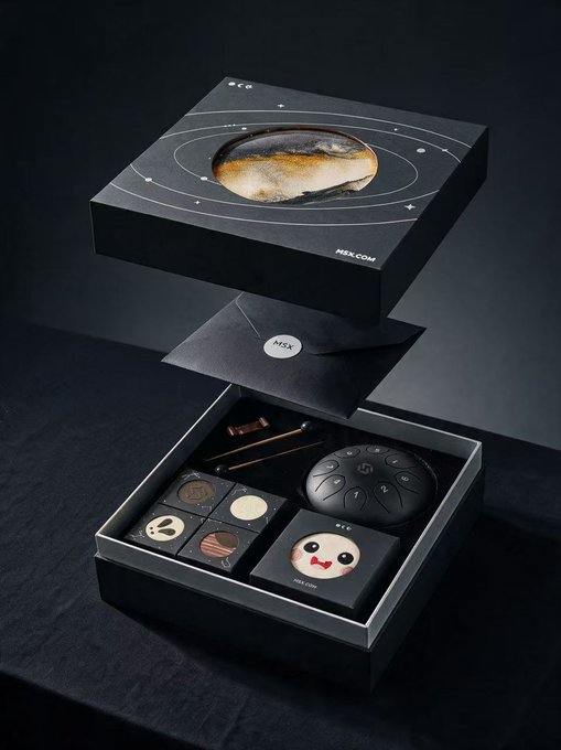 An open black MSX gift box reveals a space-themed collection featuring a planet illustration on the lid and contents including chocolate pieces, a round container with star patterns, and a small card with a cartoon face, set against a dark background.