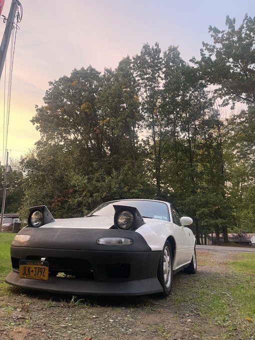 A white Mazda Miata convertible parked outdoors on grass at sunset, featuring a New York license plate reading 'ALK-3792' and pop-up headlights.