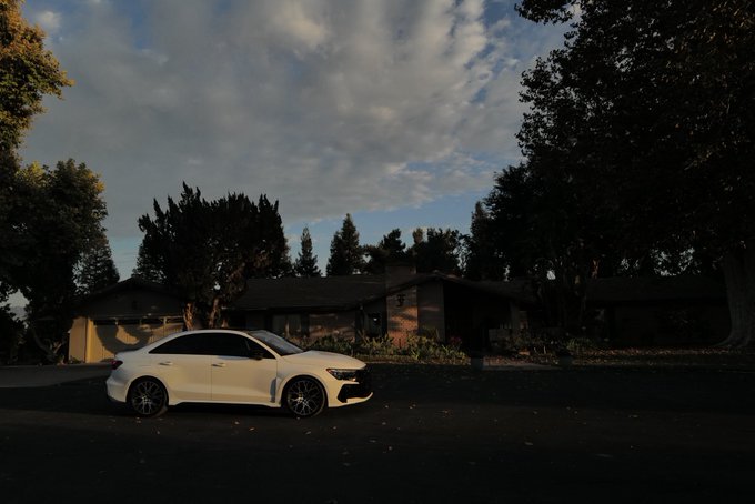A white Mercedes-Benz CLA sedan is parked in the shadow of a residential driveway at dusk, dramatically lit against a partly cloudy sky. The sleek, relatively new car ironically contrasts with the owner's sarcastic caption about already receiving an "emergency assistance unavailable" notification, suggesting the vehicle's systems are already malfunctioning not long after purchase.