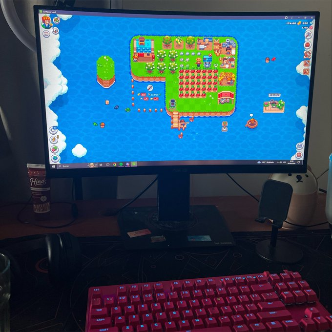 A computer monitor displays the game "Sunflower Land," showing a colorful pixel-art island with crops, trees, and buildings, alongside a red mechanical keyboard and a Pringles can. The scene illustrates the poster's morning routine of chopping trees in the Sunflower Land game before starting their day.