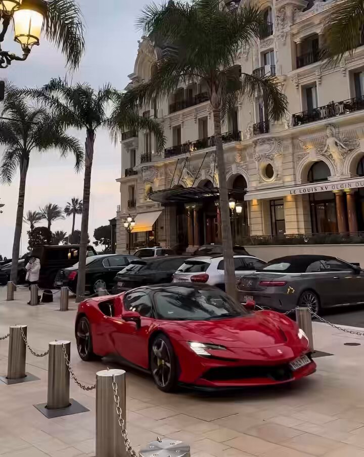 The image features a red sports car parked on a city street next to palm trees. There are several other cars parked nearby, including one that appears to be a luxury vehicle. The surrounding area includes buildings with balconies, suggesting an urban setting. A person is also visible in the scene, possibly walking by or attending to their own vehicle.