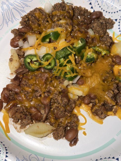 A close-up of a paper plate containing a meal with ground beef, beans, melted cheese, sliced jalapeños, onions, and shredded cheese.