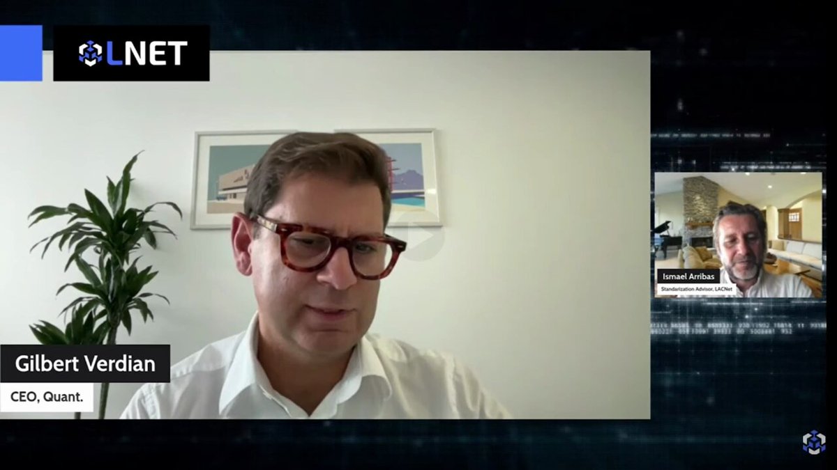 The image shows two men sitting across from each other at a computer screen during a video call. They are both wearing glasses and appear to be engaged in a conversation or presentation. The background of the image features a plant, which adds some greenery to the scene. The caption below the image reads "With @quantnetwork launching Fusion on mainnet here's a clip of CEO Gilbert Verdian which might show why this project could go mainstream Gilbert AUTHORED ISO TC307 Global Blockchain Standards which committee involved hundreds of leaders from IBM, Google, central banks," and it is written in white text.