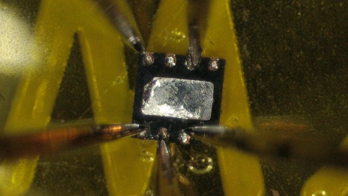 A close-up microscopic image of an electronic component showing probes making contact with solder points on a small chip.