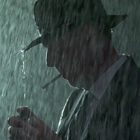 A man wearing a hat and suit smokes a cigarette while standing in the pouring rain, lit dramatically against the dark, stormy background.