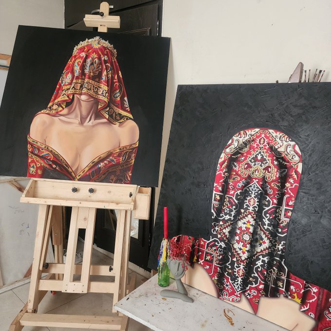 A photograph of two paintings displayed on wooden easels showing women in red patterned veils or garments, with a small decorated mannequin head and art supplies on a table in an artist's studio.
