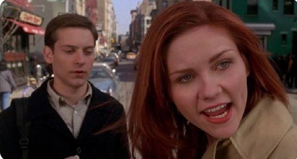 We need to write a concise 1-2 sentence caption describing what is in the image. Include any key text, numbers, tickers or names visible. The image appears to be a still from a movie (maybe "Spider-Man"? No, that's Tobey Maguire and Kirsten Dunst? Actually looks like Tobey Maguire and Kirsten Dunst in "Spider-Man" (2002). The scene: they're on a street; the woman is looking at the camera with a surprised expression. The man is looking down. There's a sign "Café" maybe "Cafe" on a red awning. Let's examine text: In background left, there's a red awning with "Cafe" maybe "Cafe" or "Café". Also maybe "Sbarro"? Not sure. Let's look: The sign is red with yellow text: "Café". It might be "Cafe" or "Café". Also