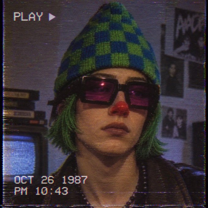 A stylized retro VHS-style portrait of a person wearing a green and blue checkered beanie, purple sunglasses, and a red clown nose. Text shows 'PLAY ▶', 'OCT 26 1987', and 'PM 10:43'.