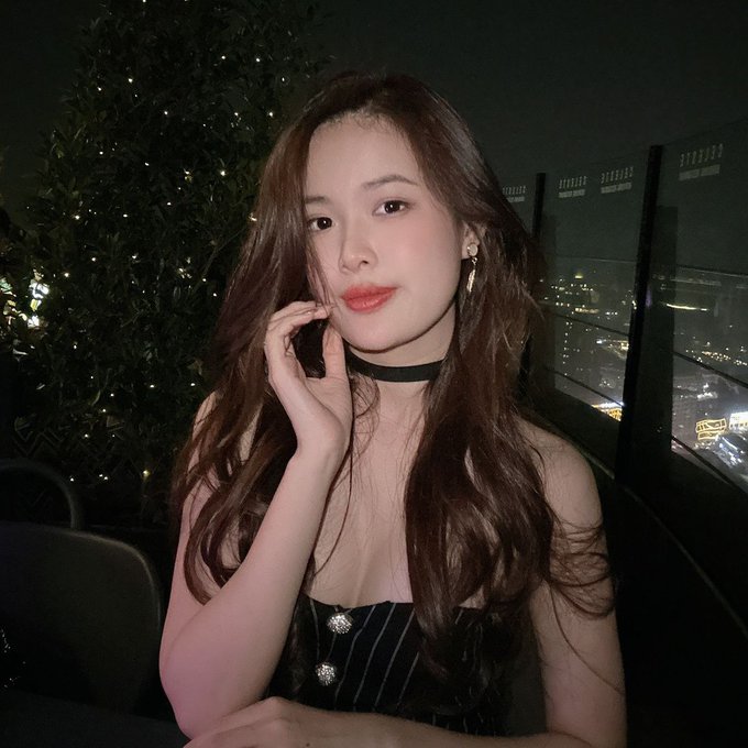 A young woman with long wavy brown hair and a black choker poses on what appears to be a balcony at night, with city lights visible in the background.