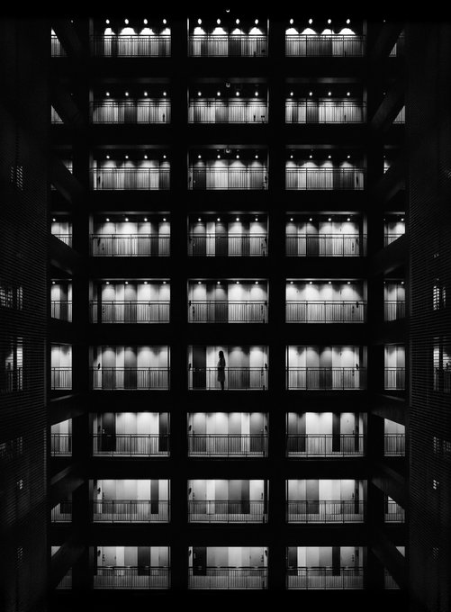 A black and white photograph looking down into a multi-story building atrium, showing rows of illuminated balconies and a lone silhouetted figure standing on one of the walkways.