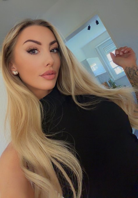 A blonde woman with full makeup, wearing a black top and a flower-shaped earring, takes a selfie indoors with natural light streaming in from windows behind her.