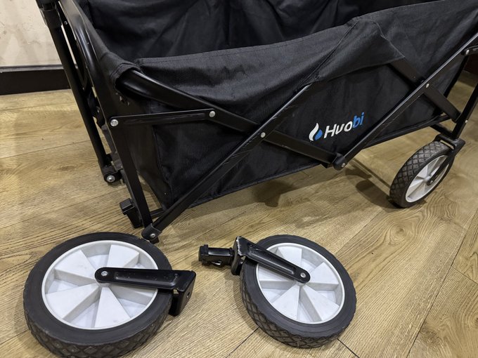 A black Huobi-branded folding wagon sits on a wooden floor with its two front wheels detached and lying beside it, appearing to have come off mid-use. The "Huobi" logo is clearly visible on the side of the wagon's fabric body, matching the post about the camper wagon gifted by the company that has now retired after years of service with the kids.