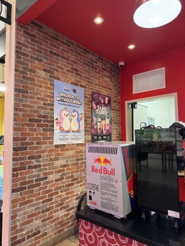A Red Bull branded mini-fridge sits on a counter inside a cafe, with 'LOOK OUT WITH US!' and 'CHIPS WITH US!' posters displayed on an exposed brick wall behind it.