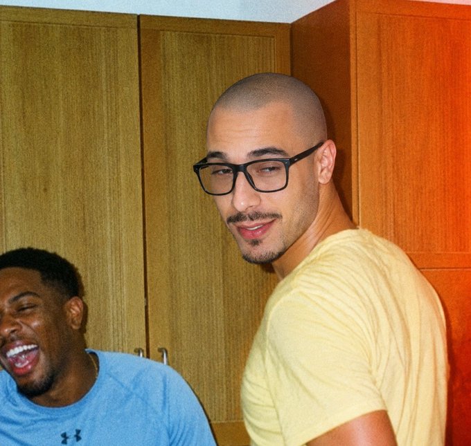The image shows two men - one with a shaved head wearing black glasses and a yellow t-shirt looking at the camera, while another man in a gray Under Armour shirt laughs in the background. The accompanying post suggests this is a moment featuring @blknoiz06, who is humbly distinguishing himself from another notable figure named Ansem, noting that while Ansem famously airdropped millions of dollars, he ("blknoiz06") only airdropped about $4.5k.