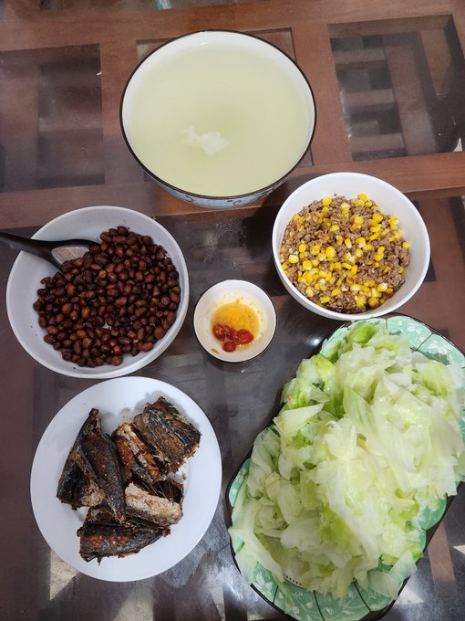 An overhead photo of a home-cooked Asian meal featuring rice porridge, soybeans, corn with minced meat, fried fish, cabbage, and a small dish of chili sauce, arranged on a glass table.