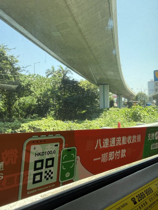 A view from inside a vehicle shows a toll payment sign in Chinese and English reading "HKD100.0" and "Welcome to use," with a road overpass and greenery visible outside.