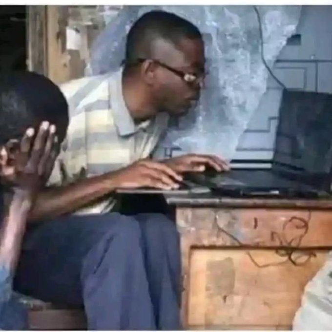 A man wearing glasses and a striped shirt sits at a wooden desk typing on a laptop, with another person partially visible beside him covering their face.