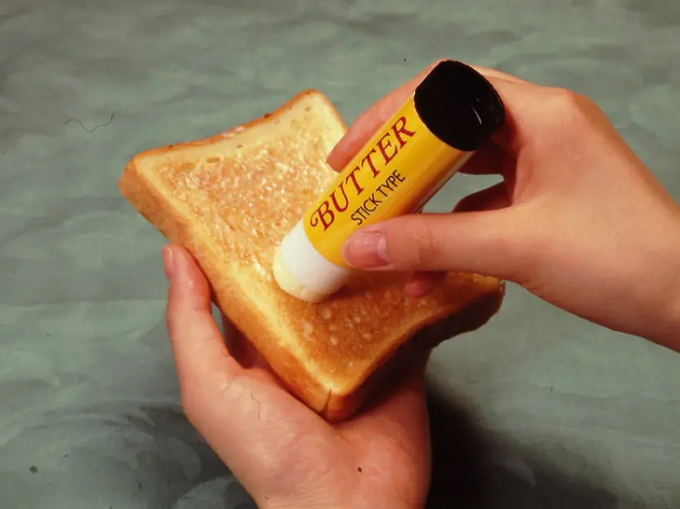 Two hands demonstrating a yellow stick-type butter product being applied to a slice of toasted bread on a green surface, with the wrapper reading 'BUTTER STICK TYPE'.