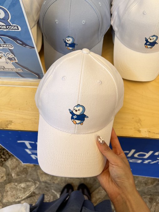 A hand holding a white baseball cap featuring an embroidered penguin mascot, with other similar caps displayed on a wooden table.