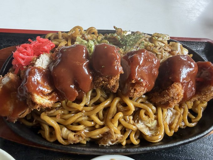 Close-up of a Japanese dish featuring breaded cutlets topped with thick brown sauce served over yakisoba noodles, accompanied by pickled red ginger.