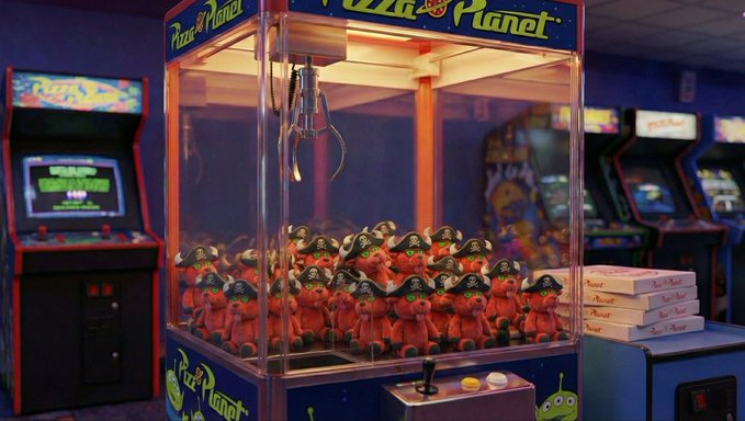 A Pizza Planet claw machine filled with plush crab toys wearing pirate hats, with classic arcade cabinets visible in the background.